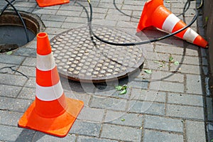 Orange cones installed around the dangerous area on the sidewalk. An open travel hatch. Laying of a new fiber-optic cable in the