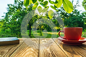 Orange cup of hot coffee and a book on a wooden table in a green garden in the morning. Summer background