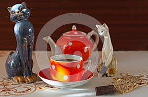 Orange cup of tea, teapot, sunflower seed cookies, chocolate and porcelain figurines of cats on white tablecloth. Breakfast or tea