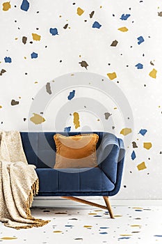 Orange cushion and blanket on blue sofa in colorful living room interior with wallpaper. Real photo