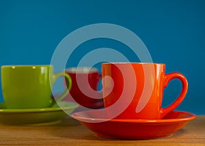 Orange, green, red cup in saucers stand on a wooden table against a blue background