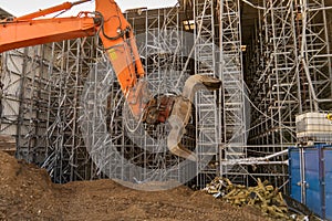 Orange gripper with excavator bucket in front of a high-bay warehouse that is being torn down