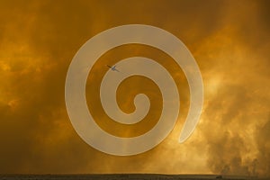 Orange hanging curtains of smoke from a large wildfire partially obscuring a firefighting plane in the desert