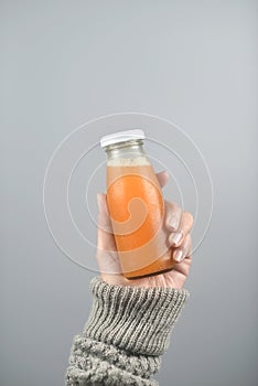 Orange juice in glass bottle on women hands with sweater on light gray background. vertical image