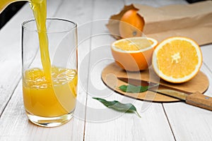Orange juice is poured into a glass on a wooden table, fresh cut oranges in the background