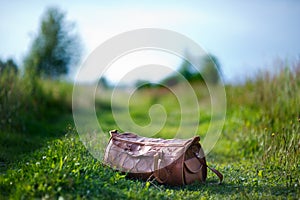 Orange leather bag on a country road on a summer evening. Green grass and vegetation around the bag