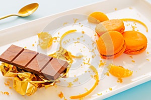 Orange macarons or macaroons on a white plate with orange peels and chocolate bar over golden paper