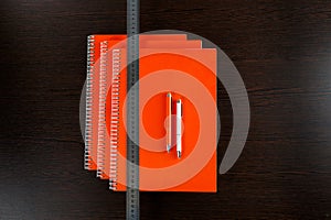 Orange notebooks lying on a dark brown wooden table with an orange and white pens and measure tape