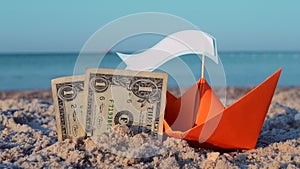 Orange paper boat, two paper dollar bills half buried in sand on sea sandy beach