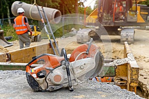 Orange petrol saw with a diamond blade for cutting concrete against a blurred background of drainage works on construction site