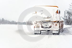 Orange plough truck on path completely covered with snow, gray sky and trees in background, view from back car driving behind -