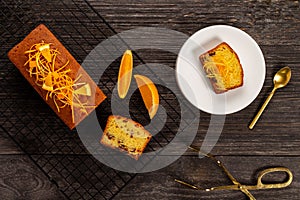 Orange pound cake on a cooling rack and slice on a plate alongside dessert tongs on dark vintage wood background, Flat lay