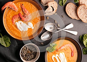 Orange pumpkin soup with shrimps, pumpkin seeds in dark bowls and bread, greenery, silver spoons