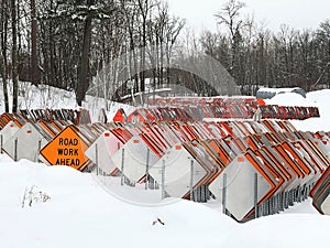 Orange ROAD WORK AHEAD sign among dozens of road signs in a snowy lot