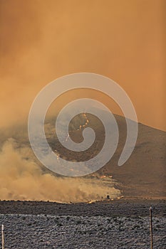 Orange smoke from a large wildfire over a partially burned hill in the desert