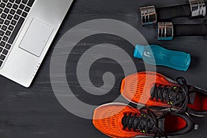 Orange sneakers, dumbbells, a water bottle and a laptop on a dark wooden background