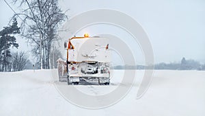 Orange snowplough truck covered with snow, on winter road, gray overcast sky background, view from behind