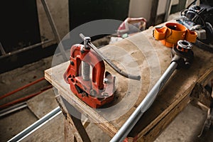 An orange steel pipe vise bolted onto a wooden makeshift table. A large ratchet is lying on the same table.