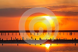 Orange sunset, dramatic and majestic sky over beach with sun reflection in water. Wood posts for salt extraction of