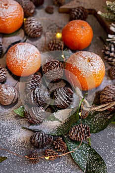 Orange tangerines on grey background in New Year`s decor with brown pine cones and green leaves. Christmas decoration with