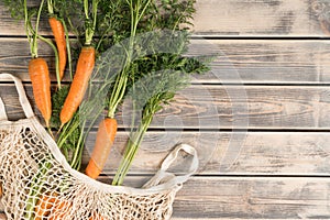 Orange tasty juicy carrot with green stem and leaves in ecological string bag on wooden background. Degradable eco bag