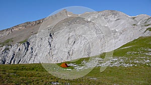 Orange tent in a remote and wild mountain landscape with green meadows and rocky peaks under a blue sky