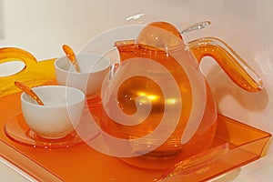 Orange transparent design teapot with two cups and plastic orange spoons on orange plastic tray.