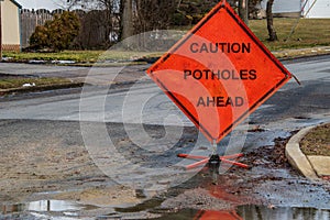 Orange triangular road sign on a small suburban street that says caution potholes ahead.