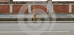 Orange waste bin hanged on yellow pole on concrete sidewalk. Brick and cement wall with erased graffiti on behind, street in front