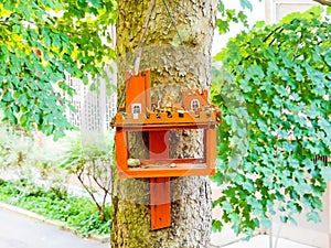 Orange wooden bird feeder with castle decor hanging on tree trunk with green foliage
