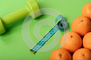 Oranges near dumbbells, measuring tape on green background, top view