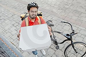 Order delivery. Smiling man with beard, in helmet, shows box of pizza to peephole