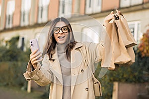 Order and pick up. Joyful fashionable woman holding a phone and shopping bags with surprised emotion on her face