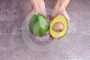 Organic avocado halves and whole fruit on the palm of a senior woman with a gray stone background. Top view. Space for text.
