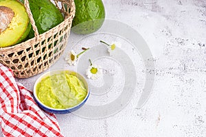 Organic avocado with seed, avocado halves in a basket, flowers, and fresh guacamole in a bowl and cloth on marble background. Top