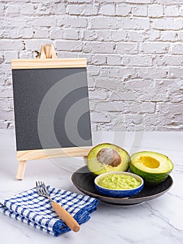 Organic avocado with seed, avocado halves, and fresh guacamole in a bowl on black plate with a small blackboard and cloth on