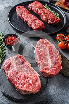 Organic feather steak steak on chopping board, marbled beef over other alternative cuts with herbs tomatoes peppercorns over grey