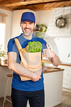 Organic food delivery service. Smiling man in blue uniform handing fresh food in a paper bag to recipient, showing okay sign.