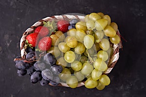 Organic fresh berries of green and blue grapes, strawberries, raspberries, blueberries in a basket on a dark concrete background.