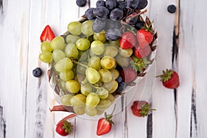 Organic fresh berries of green and blue grapes, strawberries, raspberries, blueberries in a basket on a light wooden background.
