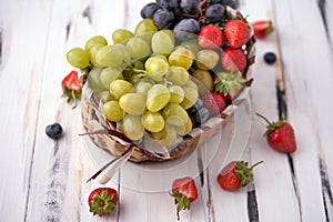 Organic fresh berries of green grapes, strawberries, raspberries, blueberries in a basket on a light wooden background.