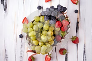 Organic fresh berries of green grapes, strawberries, raspberries, blueberries in a basket on a light wooden background.