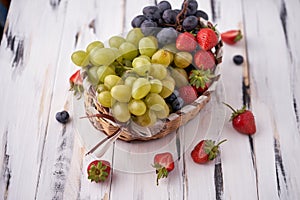 Organic fresh berries of green grapes, strawberries, raspberries, blueberries in a basket on a light wooden background.