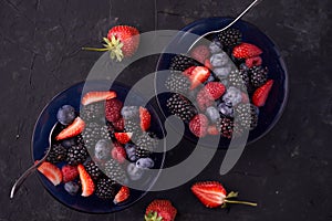Organic fresh ripe blackberries, strawberries, raspberries, blueberries in blue saucers on a dark concrete background.