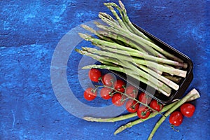Organic green asparagus and cherry tomatoes on blue textured background with copy space. Healthy dinner preparation.