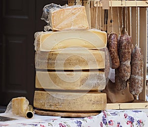 Organic hard cheese wheels stacked and salami hanging, on a shelf of an outdoor rural market in northern Italy