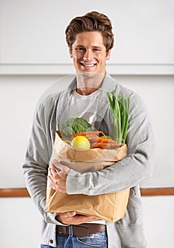 Only organic for me. A young man in a kitchen holding a brown paper bag filled with vegetables.