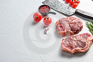 Organic top blade steak, raw beef meat with seasonings, rosemary and butcher cleaver. White textured background. Side view with