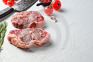 Organic top blade steak, raw beef meat with seasonings, rosemary and butcher cleaver. White textured background. Side view with