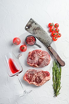 Organic top blade steak, raw beef meat with seasonings, rosemary, garnet, rose wine glass and butcher cleaver. White textured